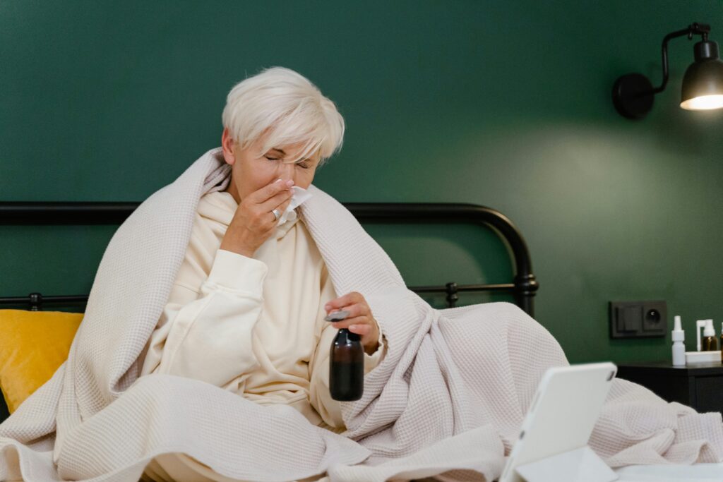 Elderly woman sits in bed feeling unwell, covered with a blanket, holding medicine.