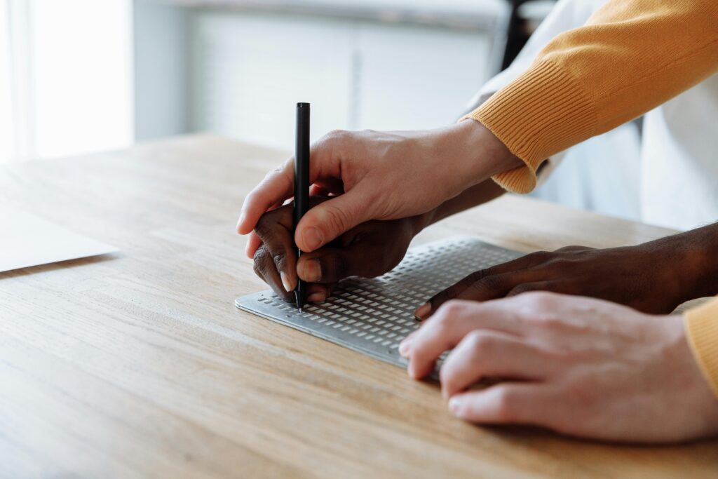 Hands skillfully guiding a student in learning braille on an embossed board.